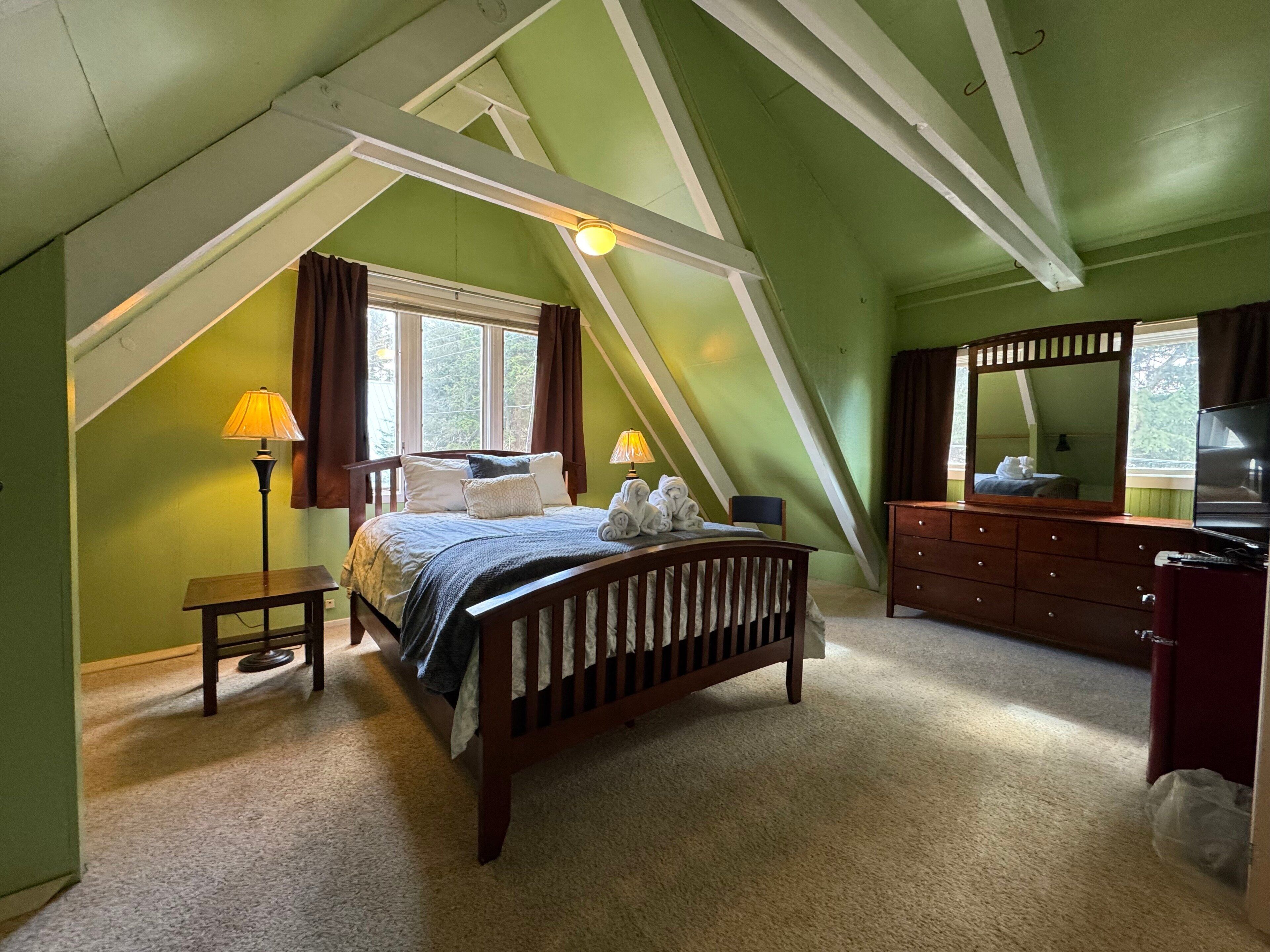 Ein Schlafzimmer mit schrägen Wänden, einem Bett, einer Kommode, einem Spiegel und einem Fenster mit Vorhängen.