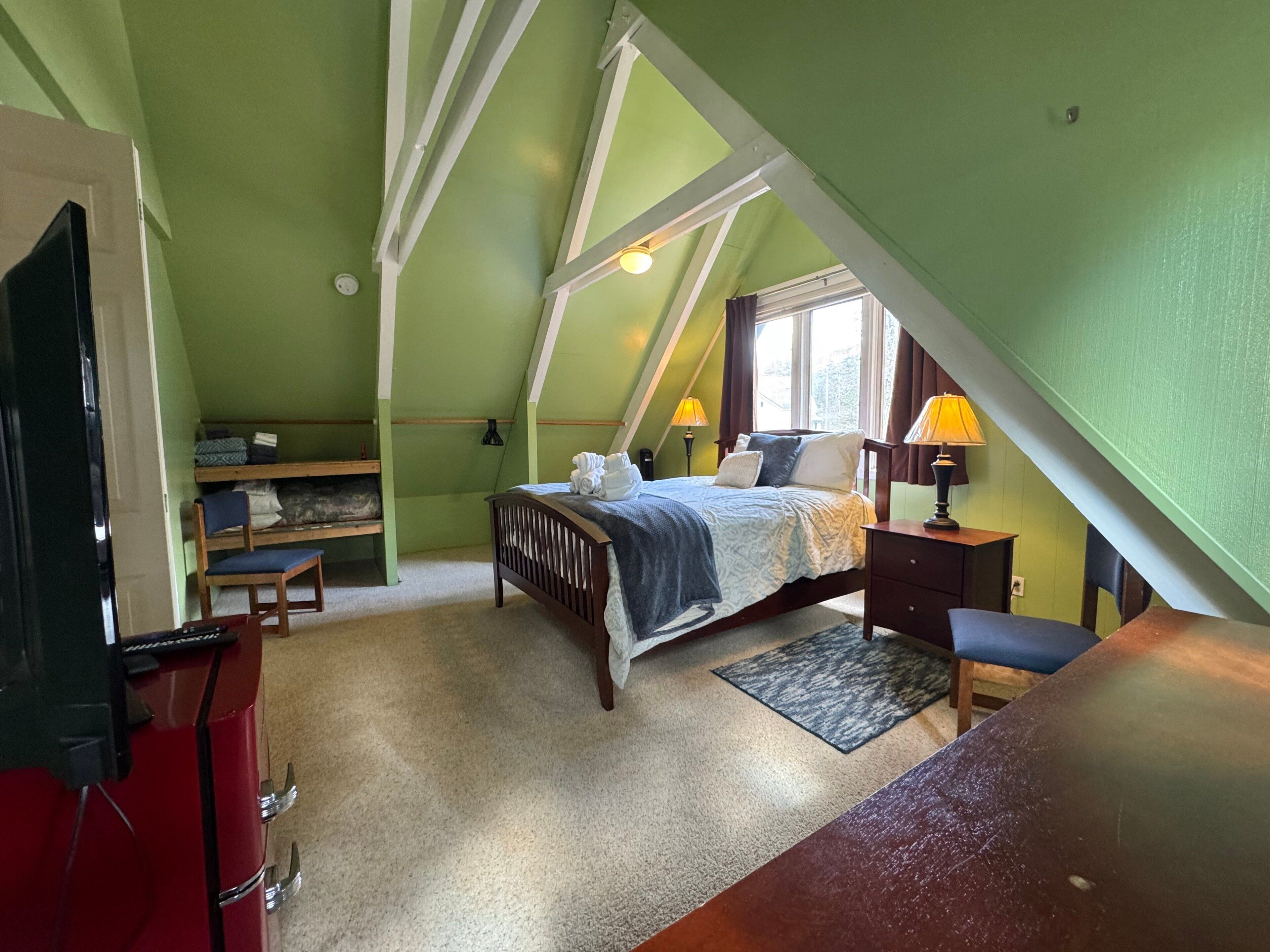 Quarto com teto inclinado, cama, escrivaninha e cadeira.