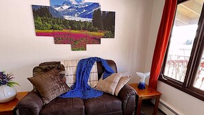 A living room with a brown leather sofa, a blue blanket, and a wall-mounted painting of a mountain landscape. - Kelli's Harbor Suites - In Auke Bay Walking Community - DISCOUNT ON TOURS (Juneau)