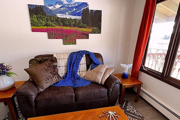A living room with a brown leather sofa, a blue blanket, and a wall-mounted painting of a mountain landscape.