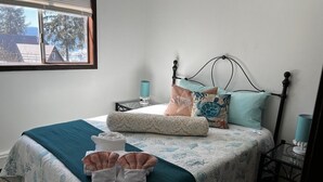 A neatly made bed with a teal bedspread and decorative pillows, a bedside table with a lamp, and a window with a view of trees and buildings. - Kelli's Harbor Suites - In Auke Bay Walking Community - DISCOUNT ON TOURS (Juneau)