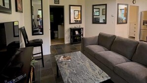 A living room with a sofa, a coffee table, and a TV. There are framed pictures on the walls and a door leading to another room.