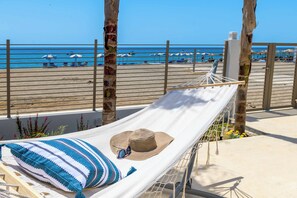 A hammock with a straw hat and striped pillow on a balcony overlooking a beach.