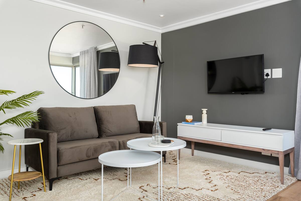 A modern living room with a grey sofa, a round mirror, a floor lamp, a TV, and a white console table.