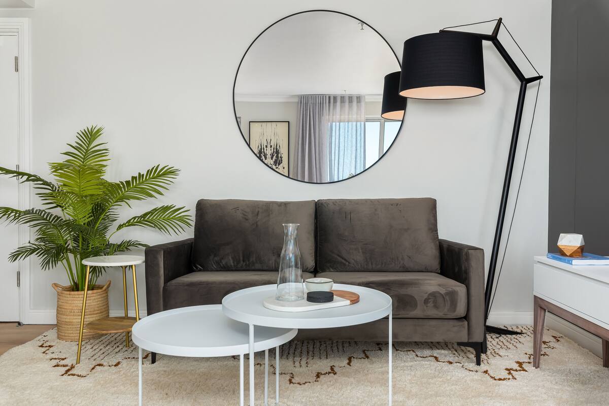 A modern living room with a grey sofa, two white round tables, a large mirror, a tall floor lamp, a potted plant, and a small side table with a vase and a candle.