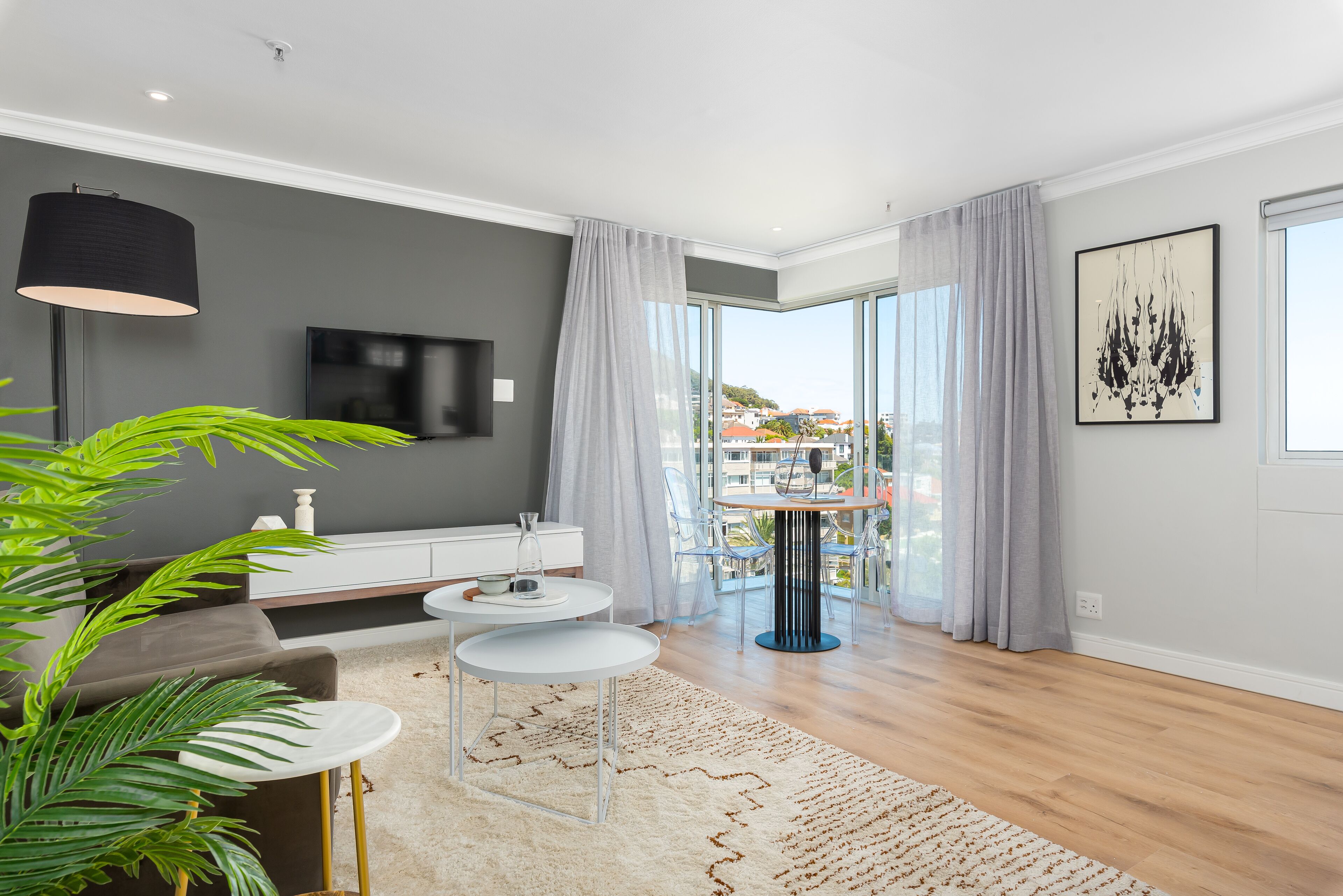 A modern living room with a grey sofa, a white coffee table, a small dining table, and a large window with a view of buildings.