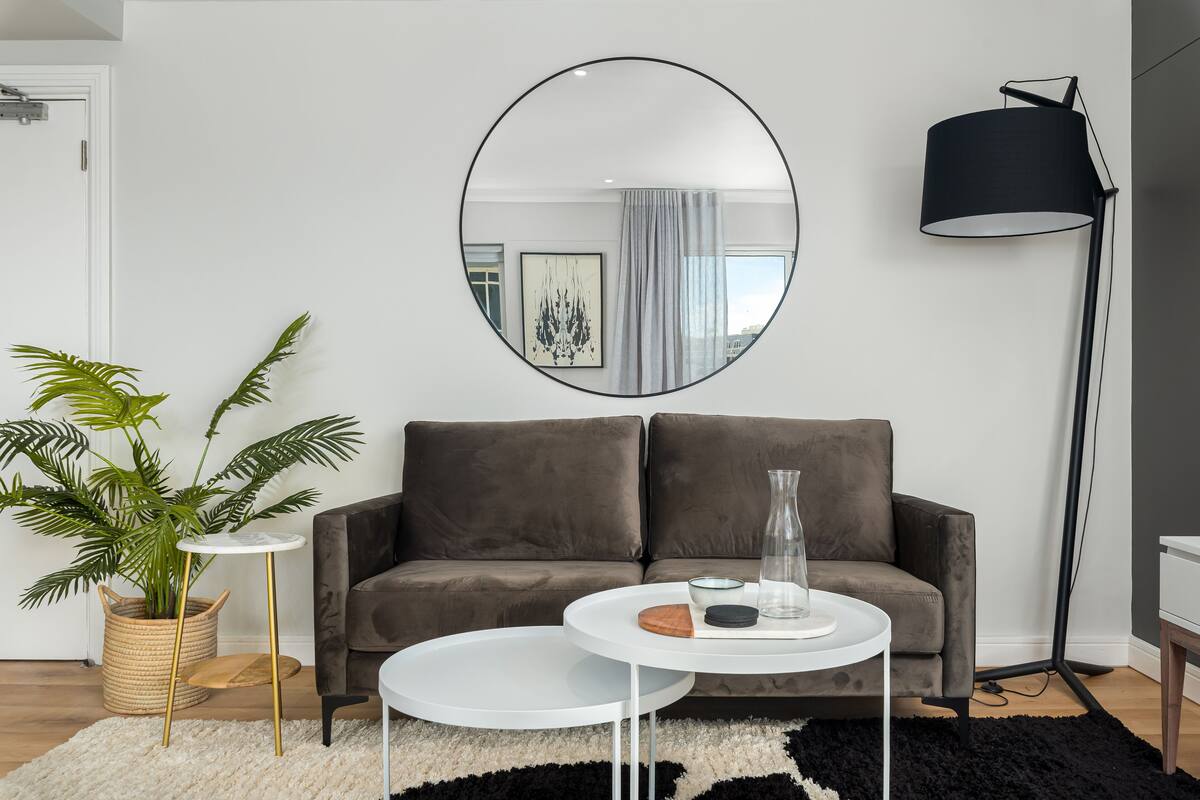 A modern living room with a grey sofa, a round mirror, a floor lamp, and a small table with decorative items.