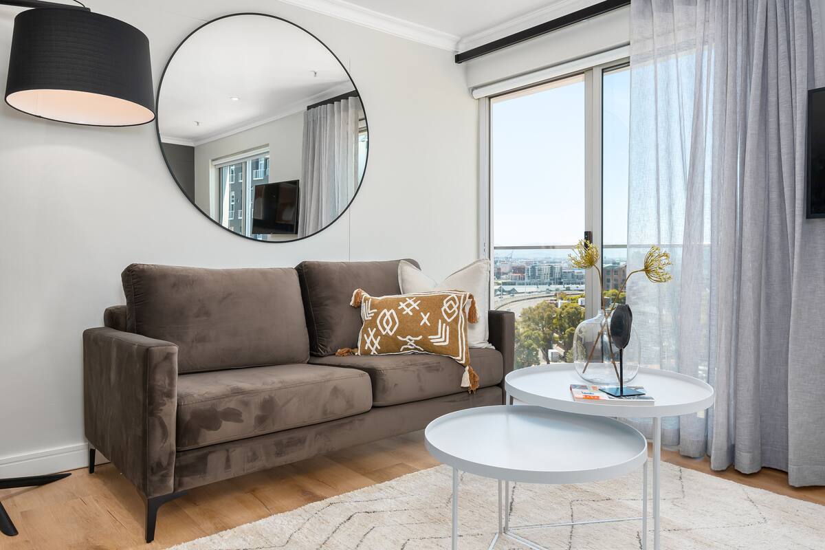 A modern living room with a grey sofa, a round mirror, a small white table, and a view of the city through large windows.