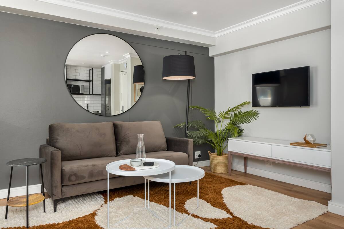 A modern living room with a grey sofa, a round mirror, a floor lamp, a TV, and a potted plant.