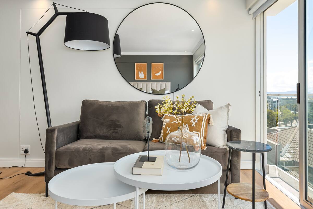 A modern living room with a grey sofa, a white coffee table, a round mirror, and a view of the city through large windows.