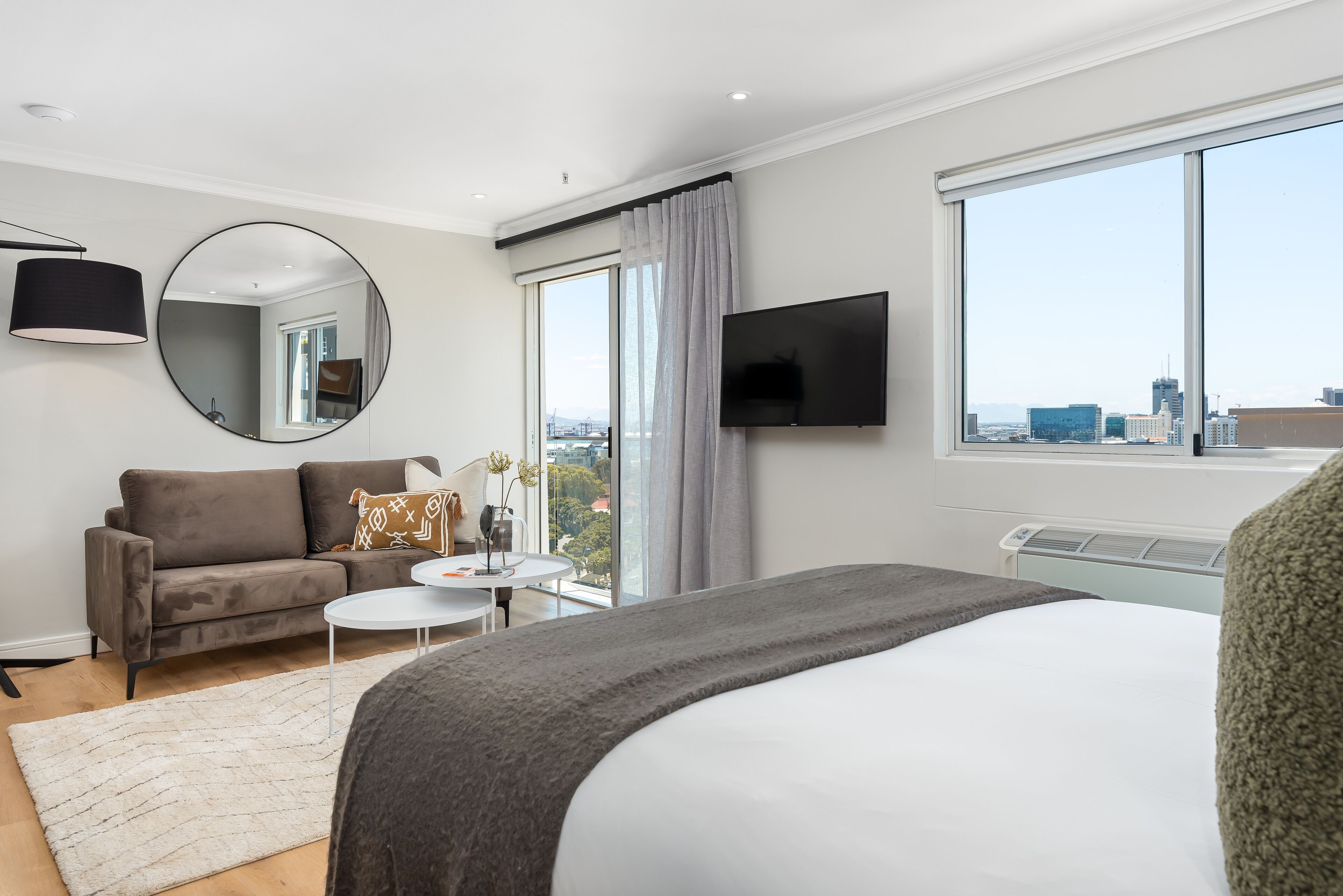 A modern hotel room with a large bed, a sofa, a small round table, a TV mounted on the wall, and a large window with a city view.