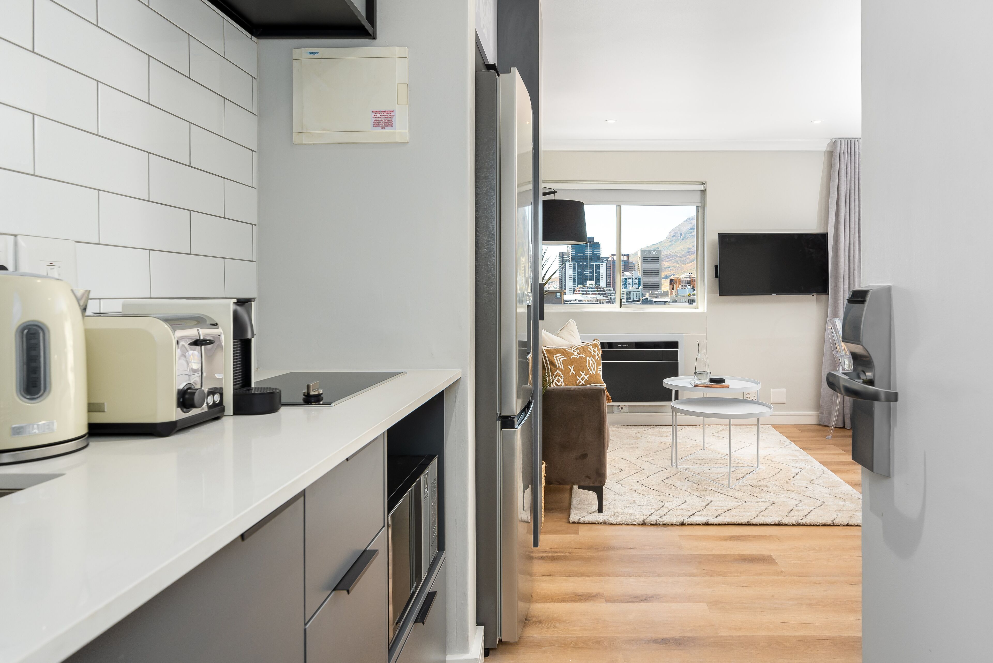 A modern kitchen with a white countertop, a refrigerator, and a toaster. There is a dining area with a table and chairs, and a view of the city through a window.