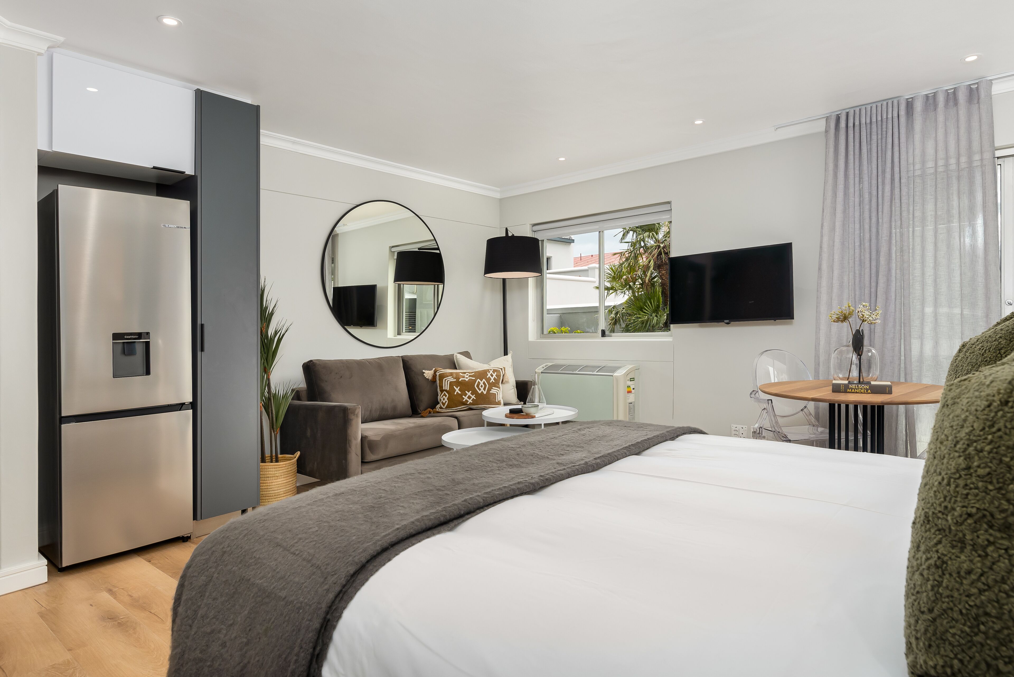 A modern hotel room with a bed, a sofa, a TV, a refrigerator, and a window with curtains.
