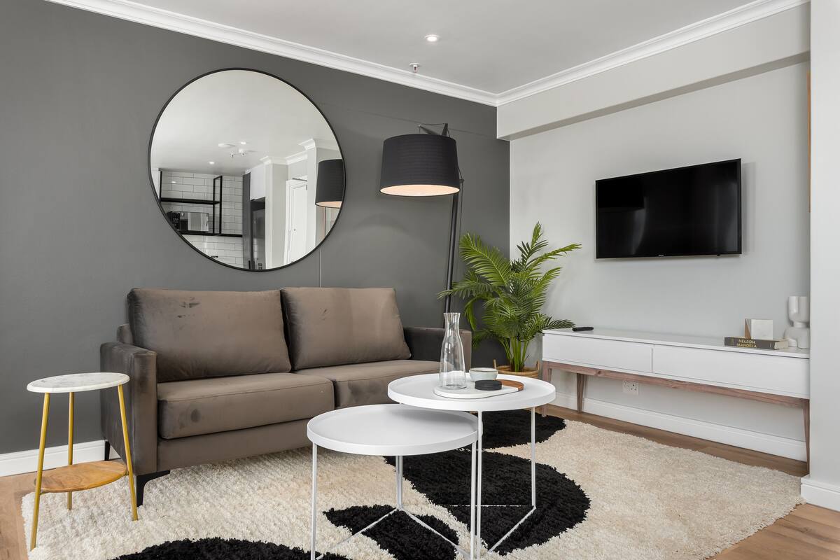A modern living room with a grey sofa, two white round tables, a large mirror, a floor lamp, a TV, and a potted plant.