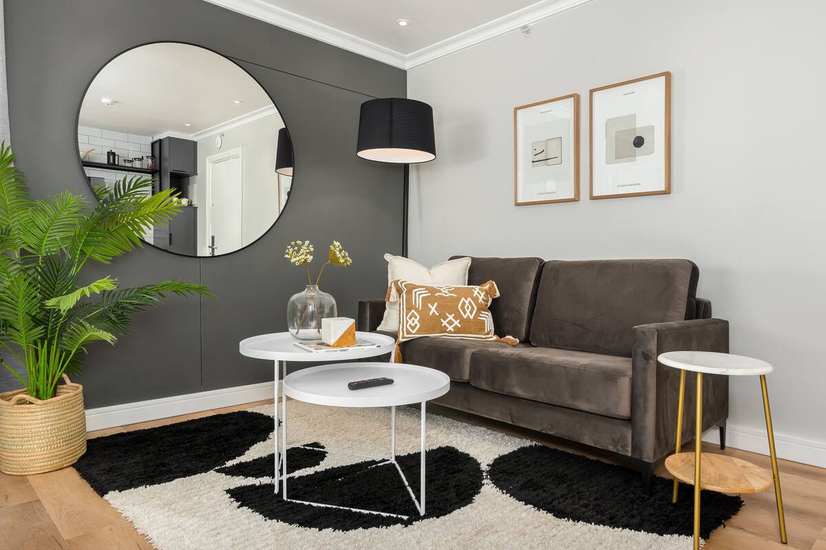 A modern living room with a grey sofa, a round mirror, a black and white rug, a small white table, and a potted plant.