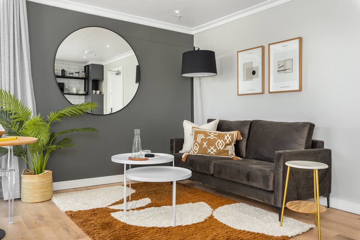 A modern living room with a grey sofa, a round mirror, a black floor lamp, and two framed artworks on the wall.