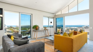 A spacious living area with a gray sofa, a yellow sofa, and a dining table with chairs. Large windows offer a view of the city and the ocean.