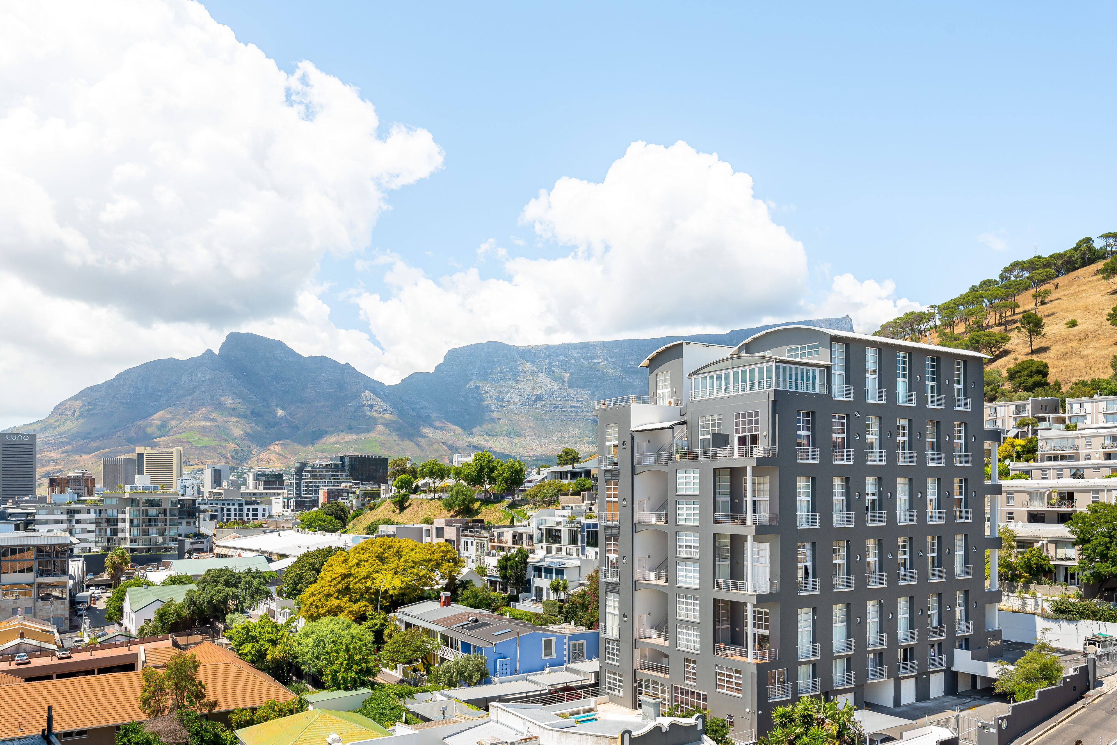 A modern multi-story building with balconies, surrounded by residential buildings and greenery, with mountains in the background.