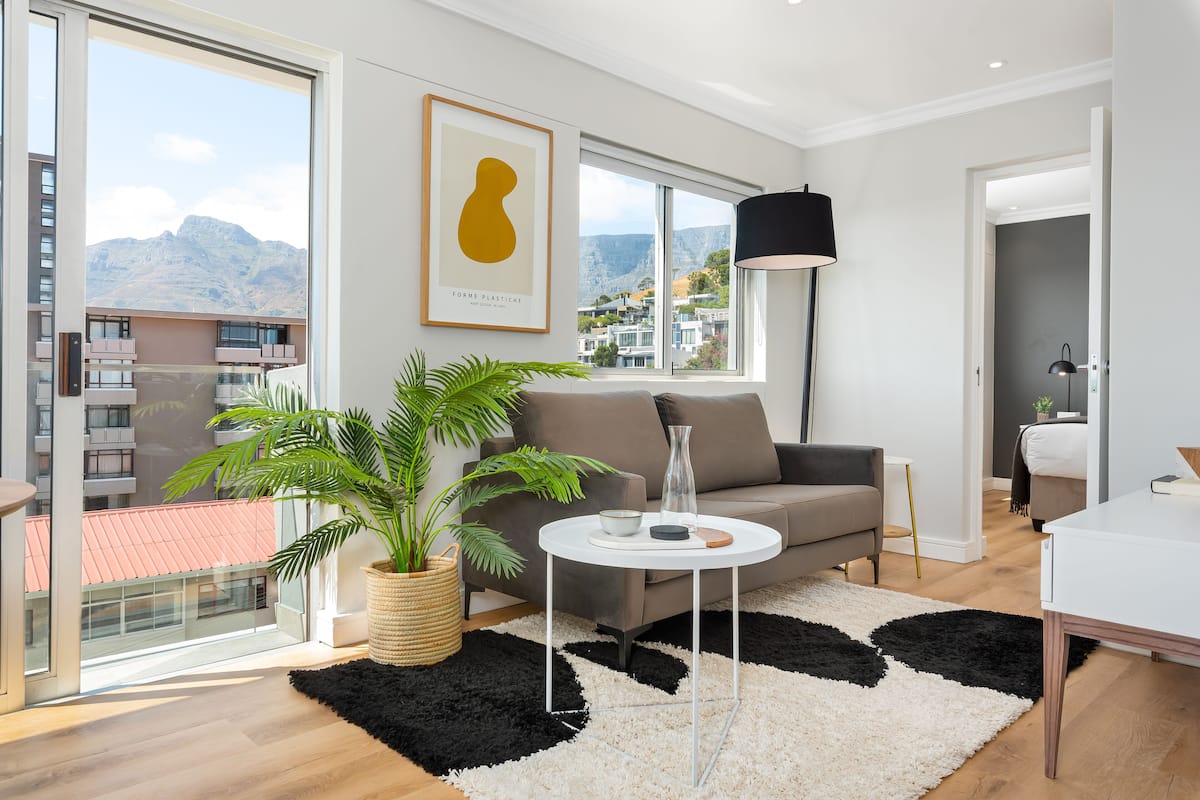 A modern living room with a sofa, a coffee table, a potted plant, and a view of mountains through large windows.