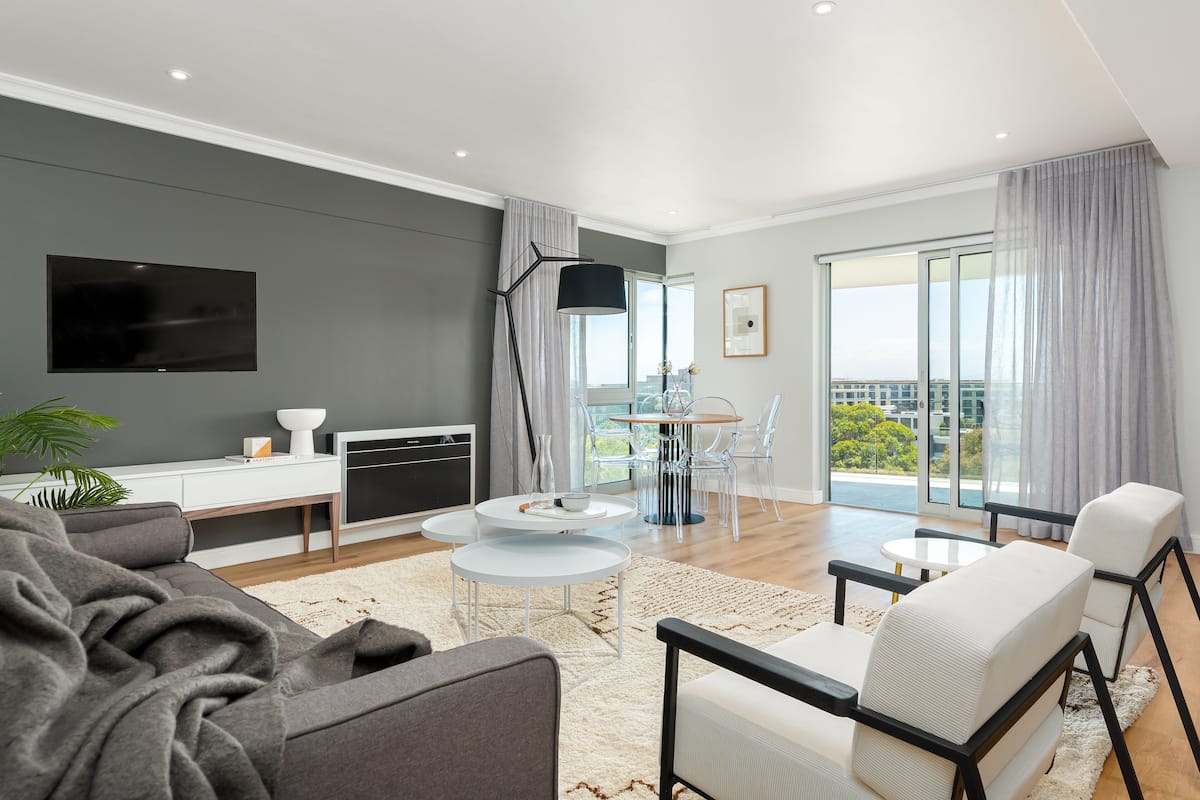 A modern living room with a flat-screen TV, grey sofa, white armchairs, and a dining area with a glass table and chairs.