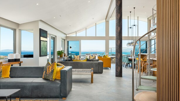 A spacious living area with a large window overlooking the sea, a staircase, and a dining area with yellow chairs.