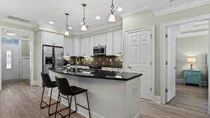 A modern kitchen with white cabinetry, a central island with bar stools, stainless steel appliances, and pendant lighting. - Golfer's Getaway Condo- Players Village TPC (Murrells Inlet)