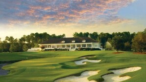 A large, white building with a symmetrical roof and multiple windows, surrounded by a well-manicured golf course with sand bunkers and a pond. - Golfer's Getaway Condo- Players Village TPC (Murrells Inlet)