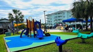 A playground with slides and climbing structures, surrounded by grass and palm trees. - Beach Vibes 1BR Condo in Litchfield (Pawleys Island)