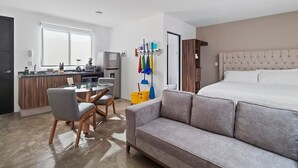 Quarto de hotel moderno com uma cama grande, área de refeições com mesa e cadeiras, cozinha compacta e banheiro.