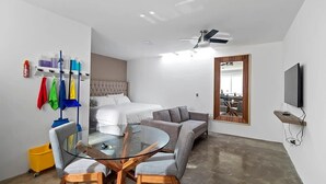 Quarto de hotel moderno com mesa de jantar de vidro, sofá, cama, ventoinha no teto e TV na parede.