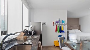 Quarto de hotel moderno com cozinha, mesa de jantar de vidro e cama com cabeceira estofada.