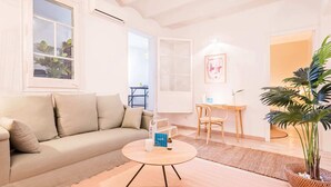 Ein Wohnzimmer mit einem beigen Sofa, einem runden Holztisch, einem kleinen Schreibtisch, einem Sessel und einer großen Kübelpflanze.