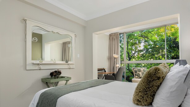 Ein Schlafzimmer mit einem großen Bett, einem Frisiertisch mit Spiegel, einem Fenster mit Blick auf Bäume und einem Schreibtisch mit Stuhl.