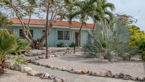 Une maison avec un toit en tuiles, entourée d’un chemin de gravier et de diverses plantes tropicales.