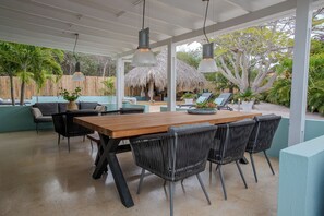A covered outdoor dining area with a wooden table and black wicker chairs, surrounded by a thatched-roof structure and lush greenery. - Villa Paradis with a private pool, palapa, tropical garden, and complete privacy (Grote Berg)