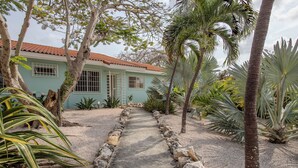 Un chemin menant à une maison dont le toit est recouvert de tuiles rouges, entourée de plantes tropicales et bordée d’un sentier de gravier.