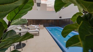 A rooftop pool area with lounge chairs and tables, surrounded by large green plants.