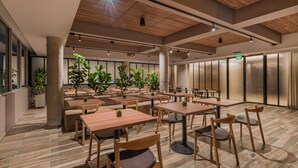 A spacious indoor dining area with multiple wooden tables and chairs, large windows, and potted plants.