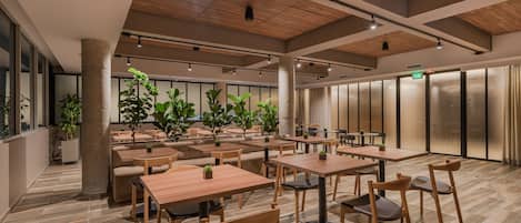 A spacious indoor dining area with multiple wooden tables and chairs, large windows, and potted plants.