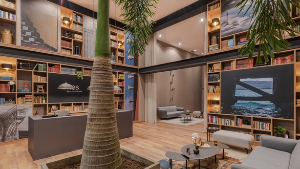 A spacious library with multiple bookshelves, a central seating area with a coffee table, and a large window with a view of the sea.
