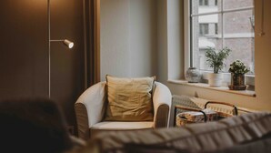 Una habitación acogedora con un sillón blanco curvo, una lámpara de pie, una ventana con plantas en macetas y vistas a edificios.