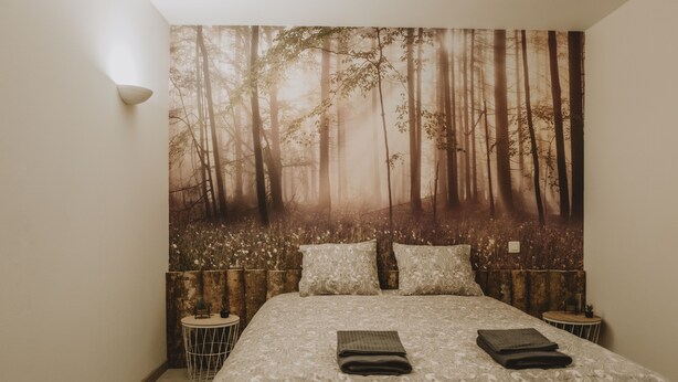 Un dormitorio con un gran tapiz de temática forestal en la pared, una cama bien hecha con dos almohadas y dos mesitas de noche con lámparas.