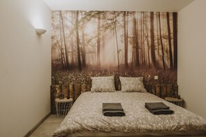 Une chambre à coucher avec une grande tapisserie murale sur le thème de la forêt, un lit bien fait avec deux oreillers et deux tables de chevet avec des lampes.