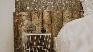 Esquina de una habitación con pared de madera rústica, una pequeña mesa blanca de metal con una planta en maceta, y una cama con colcha estampada.