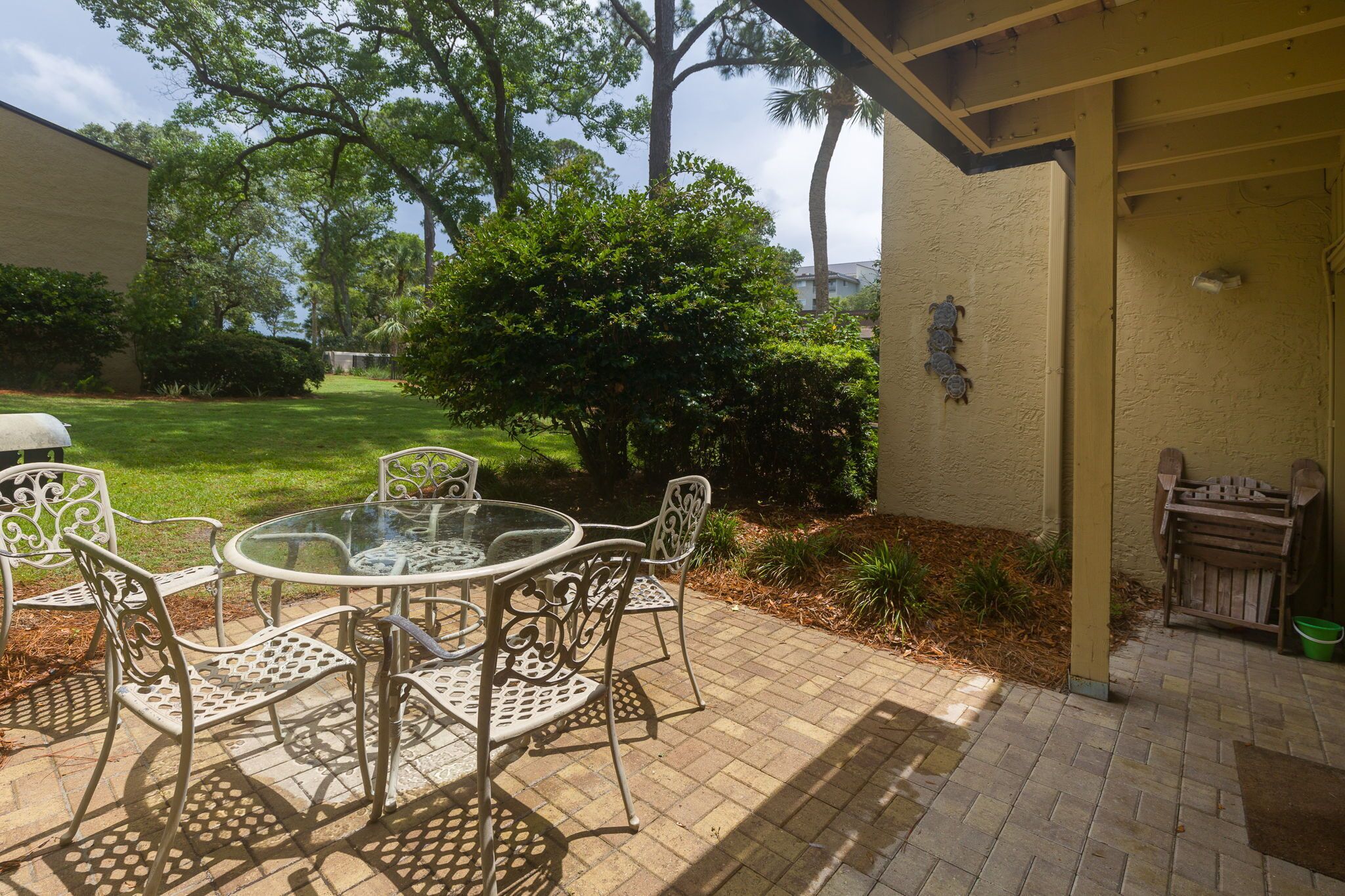 An outdoor patio with a round glass table and white metal chairs, surrounded by a brick floor and greenery.