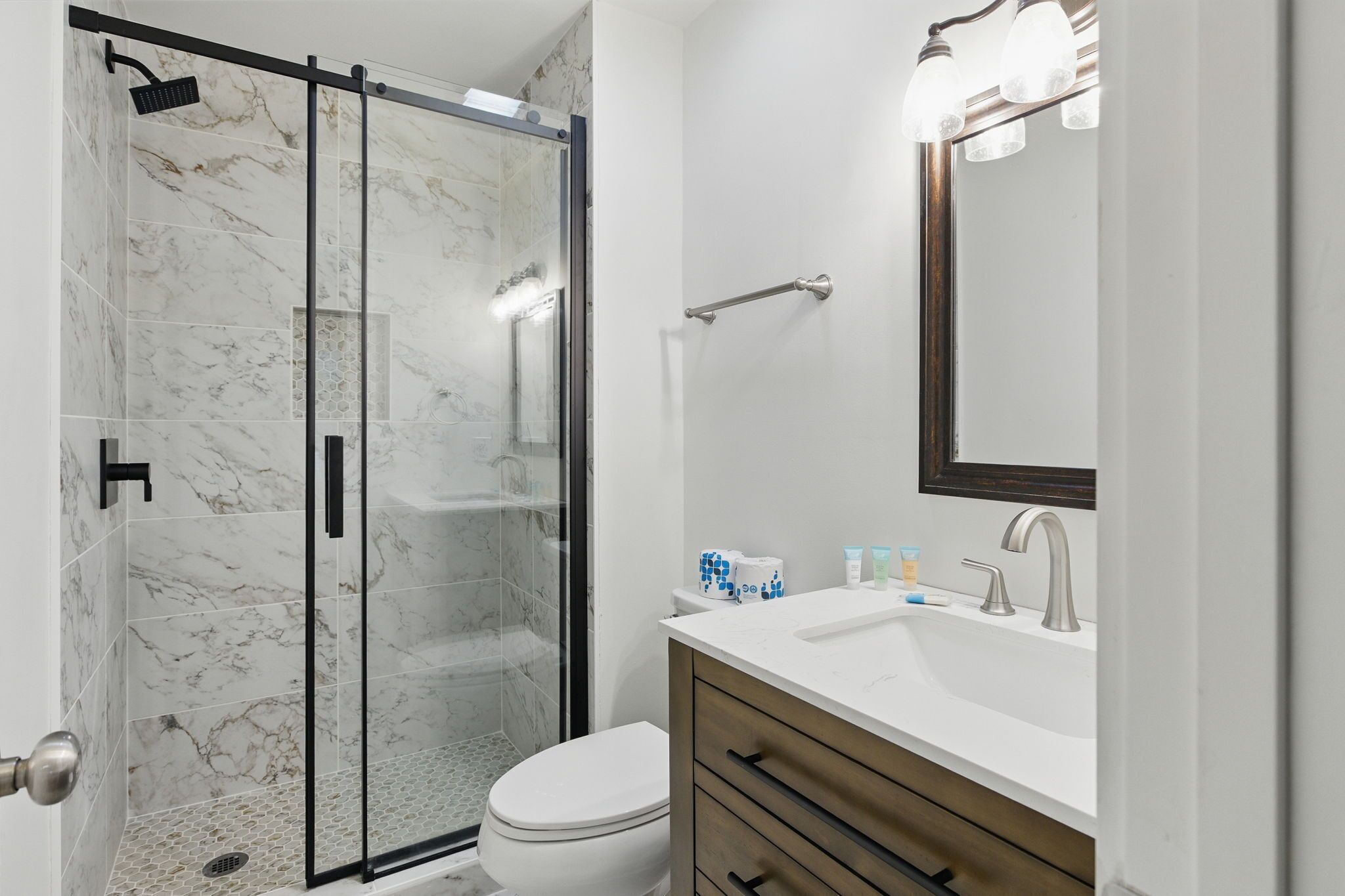 A modern bathroom with a glass-enclosed shower, a white toilet, a vanity with a mirror, and toiletries.