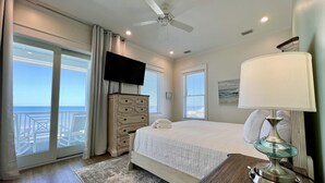 A bedroom with a bed, a dresser, a desk with a lamp, a TV, and a view of the ocean.