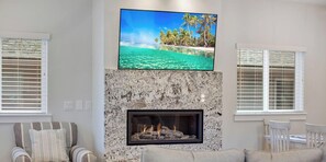 A living room with a fireplace, a television mounted above it, and a view of a beach through the window.