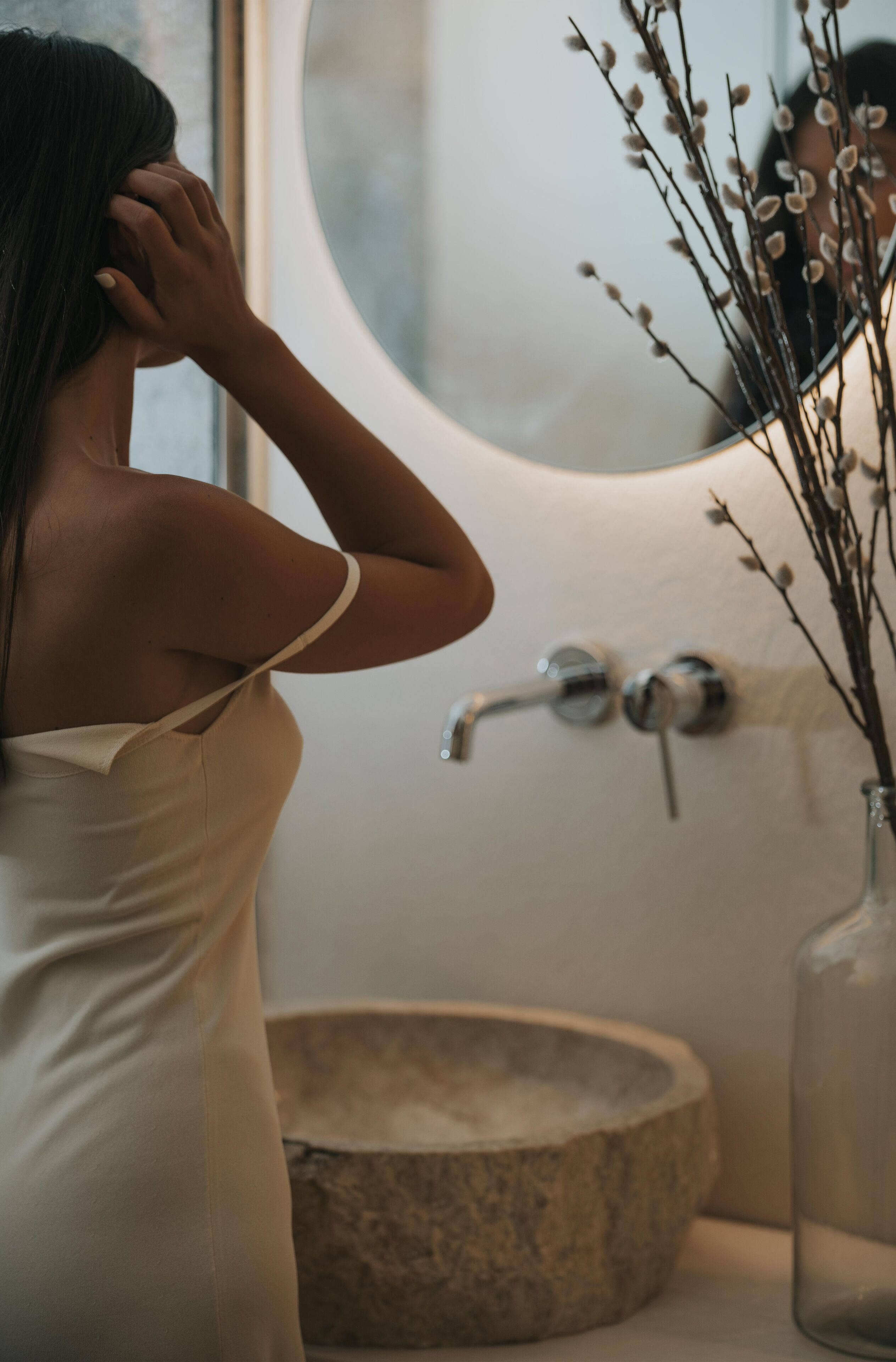 A person in a white dress standing by a bathroom sink with a stone basin and a vase with branches.