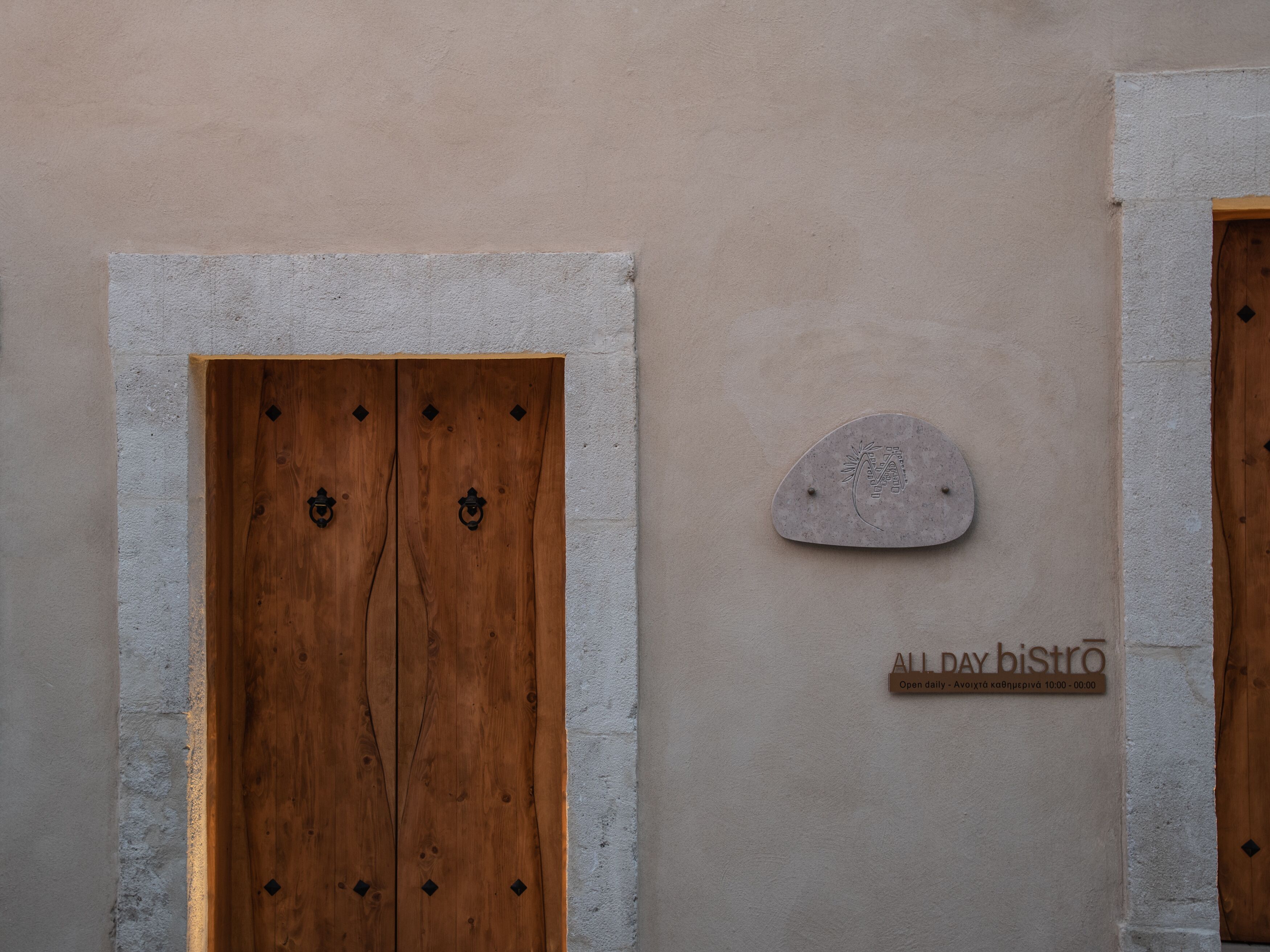 Una porta de fusta amb una placa de pedra i un rètol que indica 'ALLDAY bistro' amb l'horari d'obertura.